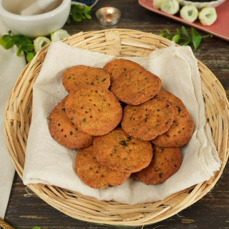 Basket of round mathris on a wooden table with a candle and small bowl in the background.